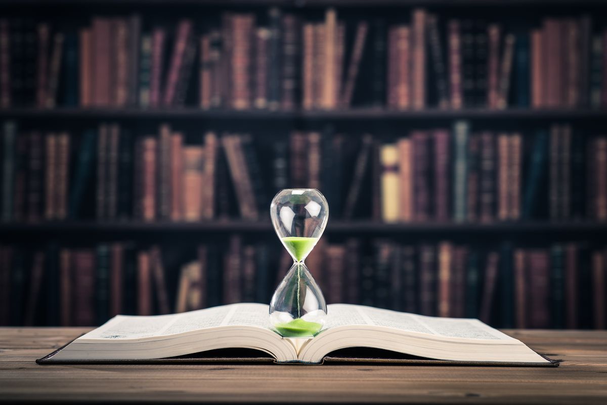 An hourglass standing on an open book in front of bookshelves