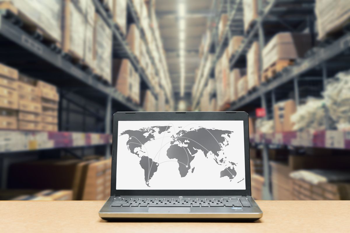 A laptop on a wooden desk showing a world map with a blurred warehouse behind it.