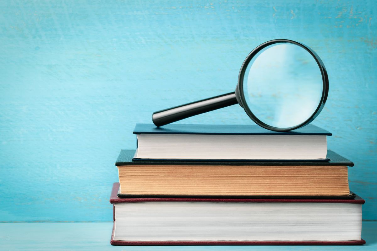 Three hardcover books stacked beneath a magnifying glass against a teal wall