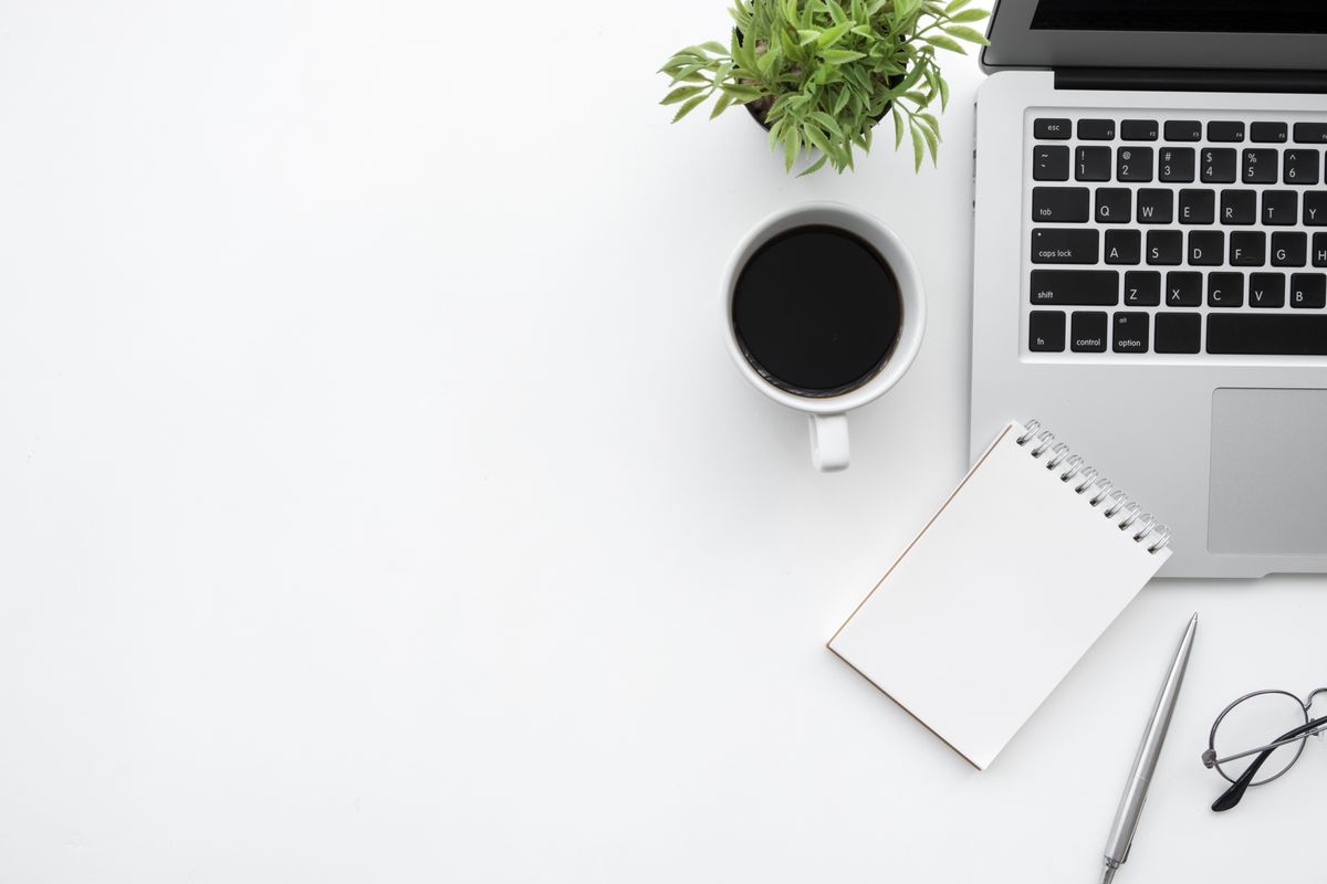An overhead view of a laptop, a cup of coffee and a blank notebook on a white desk.